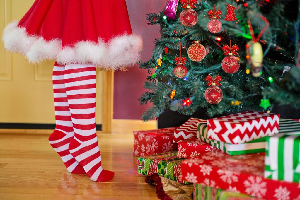 An elf's legs are seen near a holiday tree with gifts